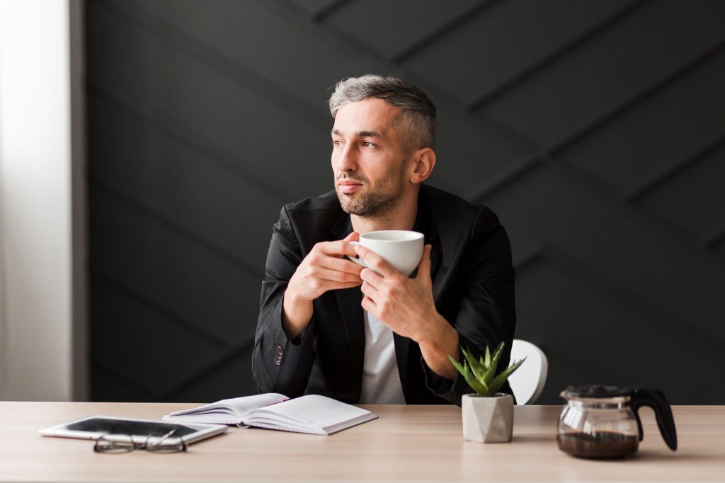Young professional man under 30 with premature grey hair wearing black jacket, holding coffee mug at desk